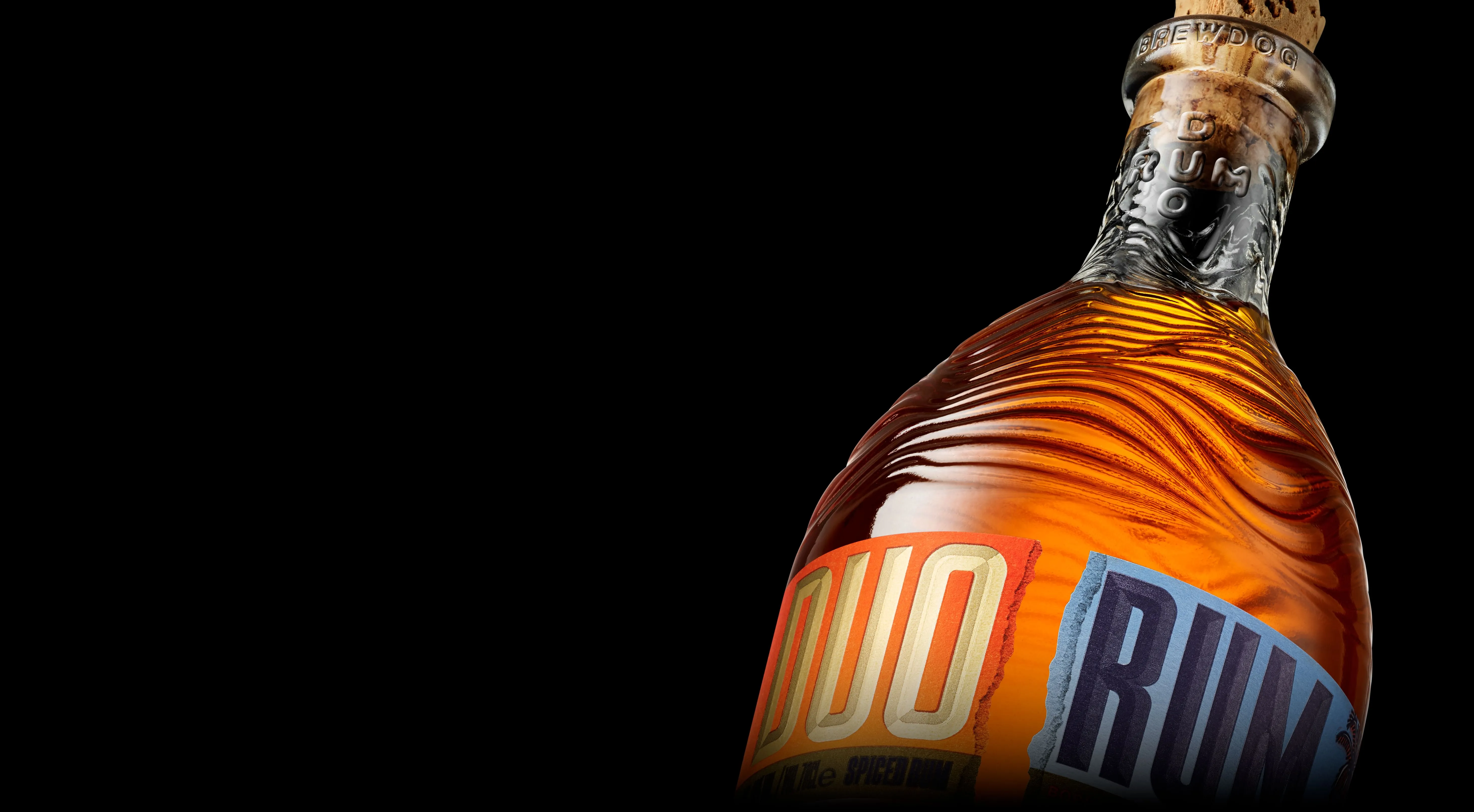 Close up of Brewdog's Duo Rum on a black background. The glass is uncoloured, showing rich amber rum inside. The shoulder of the bottle has sweeping waves of texture. The neck has an overlaid DUO RUM detail and top of the bottle is corked.