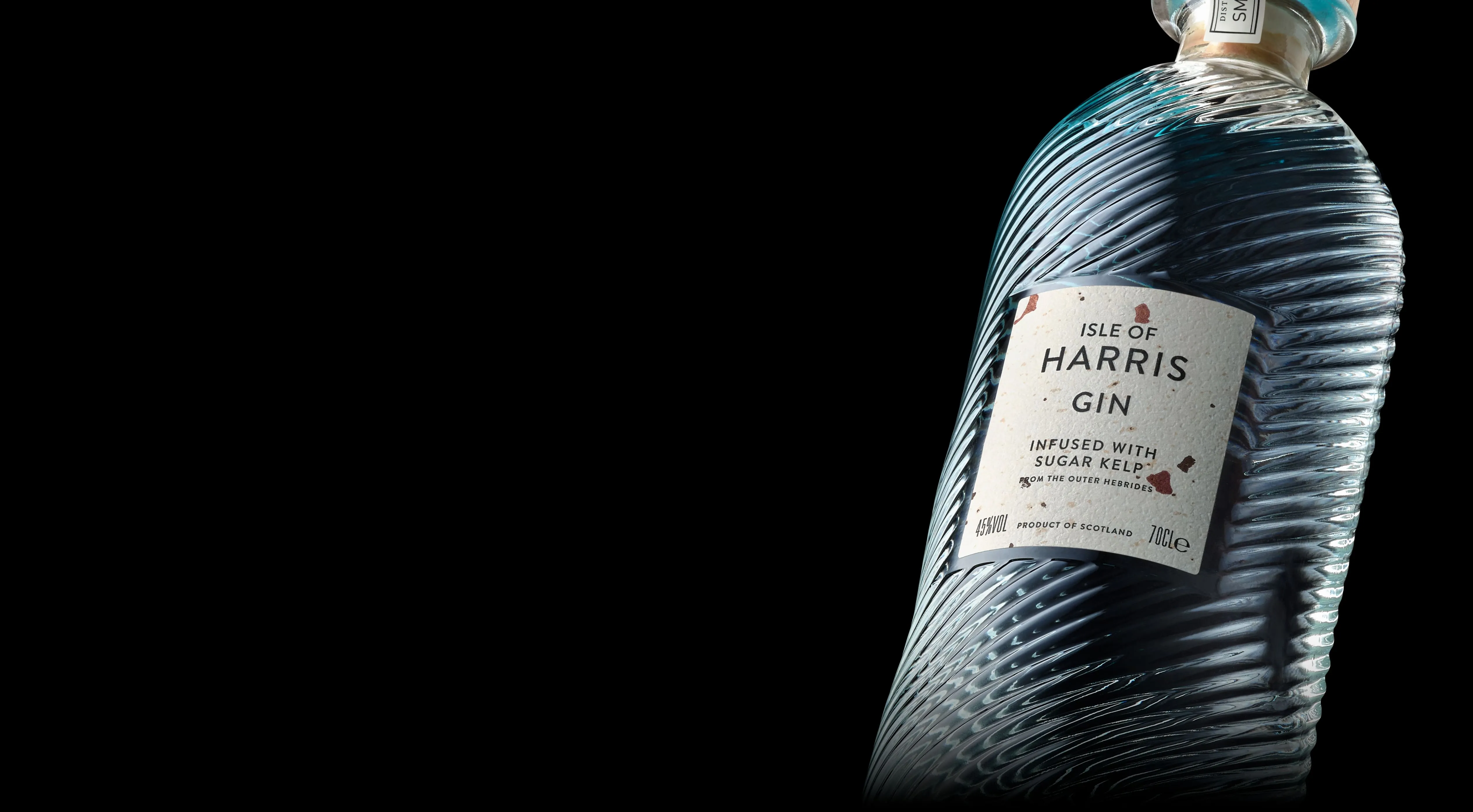 Close up of Harris Gin bottle on a black background, the glass appears light blue. The bottle is covered in a unique spiral texture. The label on the front is lightly textured and sparsely flecked with colour. It reads Isle of Harris Gin, infused with sugar kelp from the outer hebrides.