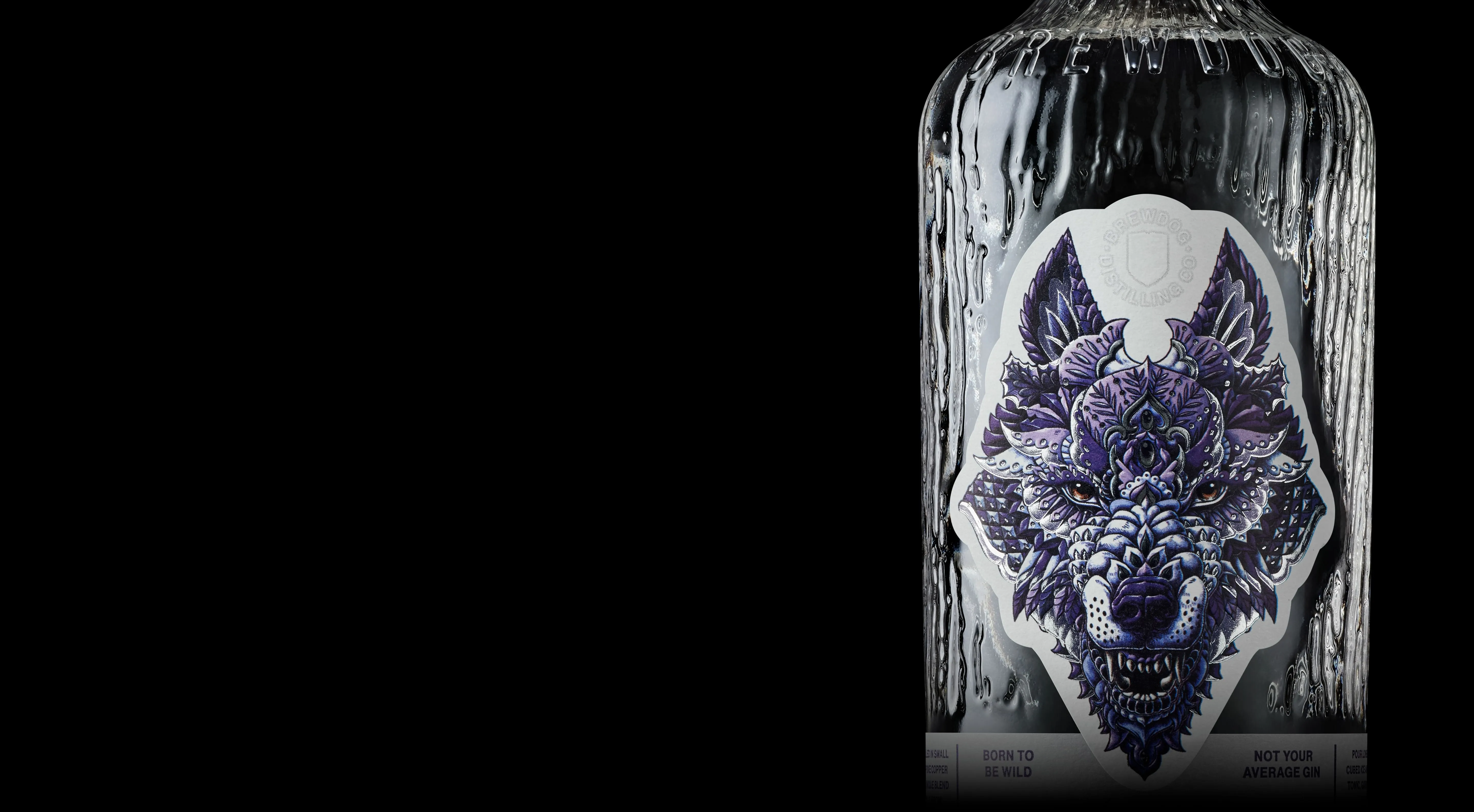 Close up of Brewdog Lonewolf bottle. A clear glass bottle with sparse texture all over. The label on the front is intricate with white and purple floral shapes building up the face of a wolf